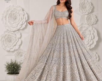 Puede incluir: Un conjunto de lehenga choli plateado que incluye un top corto sin mangas y una falda voluminosa hasta el suelo, ambos decorados con intrincados bordados plateados, lentejuelas y espejos, junto con una dupatta transparente a juego.