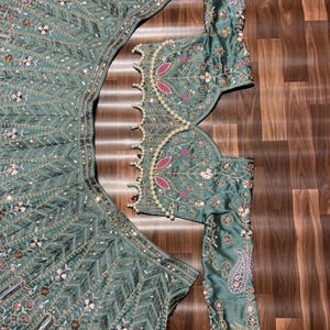Puede incluir: Un conjunto de lehenga verde salvia que incluye una blusa de manga larga y una falda acampanada. Ambas piezas est&aacute;n adornadas con intrincados bordados plateados, lentejuelas y peque&ntilde;os detalles florales rosas. El borde tiene perlas blancas.