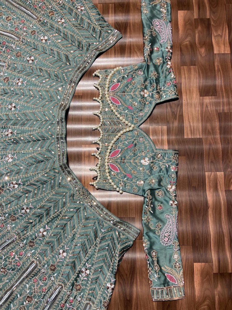 Puede incluir: Un conjunto de lehenga verde salvia que incluye una blusa de manga larga y una falda acampanada. Ambas piezas est&aacute;n adornadas con intrincados bordados plateados, lentejuelas y peque&ntilde;os detalles florales rosas. El borde tiene perlas blancas.