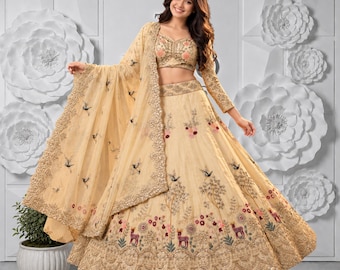 Puede incluir: Un conjunto de lehenga choli en color beige que incluye una blusa sin mangas y una falda voluminosa hasta el suelo. El conjunto está decorado con bordados intrincados de pájaros, motivos florales y ciervos, con detalles en lentejuelas doradas.