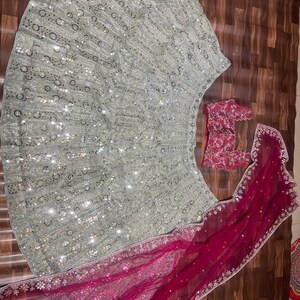 May include: A light green lehenga skirt featuring intricate silver embroidery and sequins, paired with a dark pink sleeveless blouse and a matching dark pink sheer dupatta with silver embroidered borders and scattered sequin embellishments.