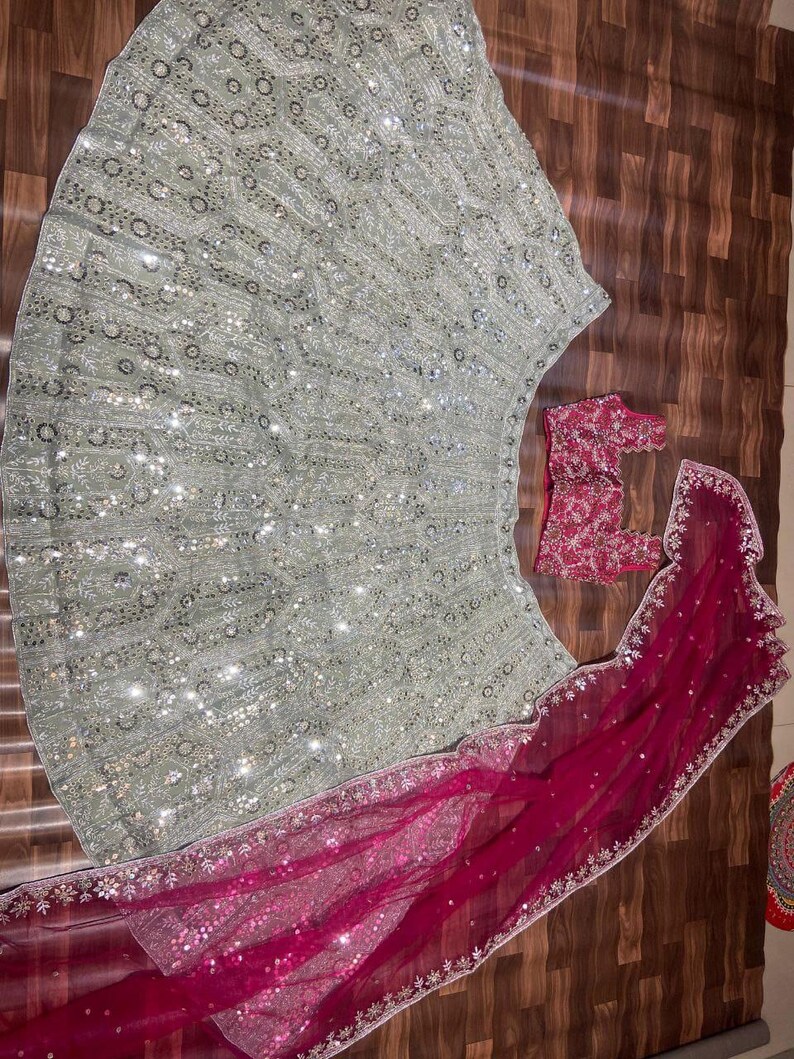 May include: A light green lehenga skirt featuring intricate silver embroidery and sequins, paired with a dark pink sleeveless blouse and a matching dark pink sheer dupatta with silver embroidered borders and scattered sequin embellishments.