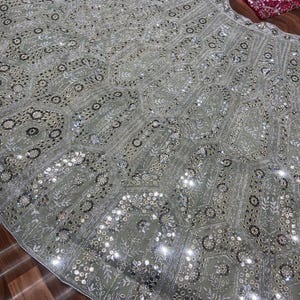 May include: A light sage green lehenga skirt featuring an intricate geometric pattern of silver embroidery, sequins, and small mirrors. A separate dark magenta blouse with dense silver embroidery and mirror work is positioned at the top corner.