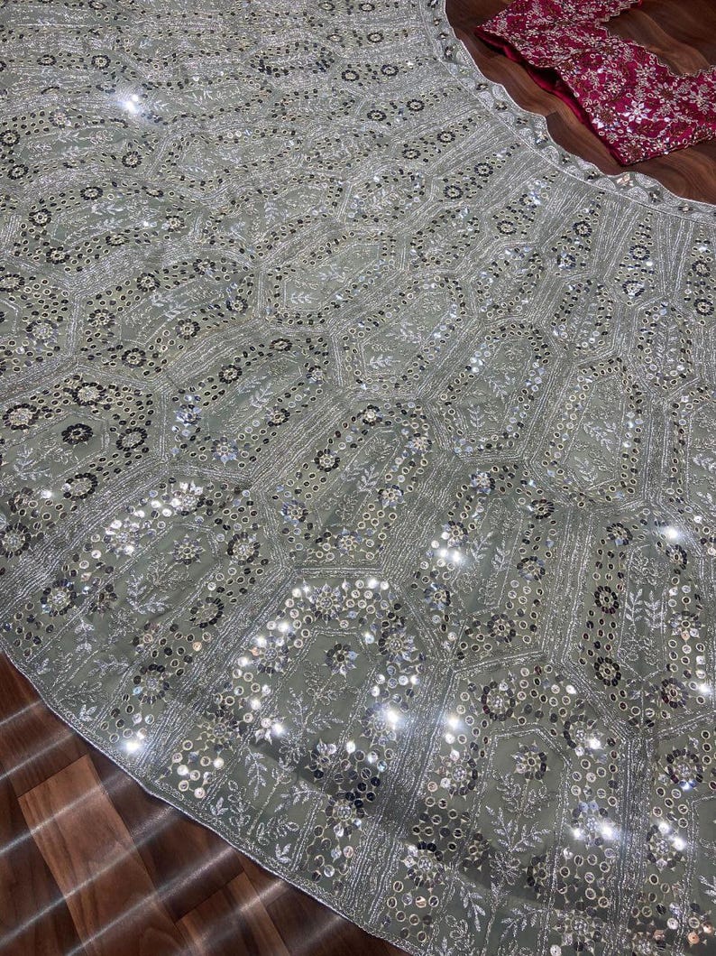 May include: A light sage green lehenga skirt featuring an intricate geometric pattern of silver embroidery, sequins, and small mirrors. A separate dark magenta blouse with dense silver embroidery and mirror work is positioned at the top corner.