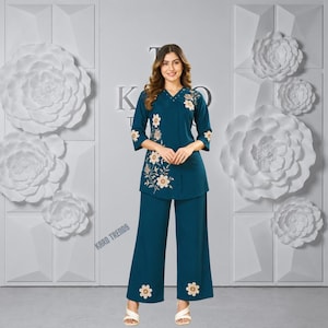 May include: A teal two-piece set consisting of a three-quarter sleeve tunic and wide-leg trousers. The tunic features a V-neckline and cream-coloured floral embroidery on the front and sleeves. The trousers have matching floral embroidery at the hems.