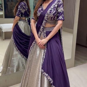 Puede incluir: Un conjunto de lehenga en color morado intenso y plata metalizada que incluye una blusa de manga corta con intrincados bordados plateados, una falda de talle alto a juego y un dupatta morado drapeado con un borde bordado en plata.