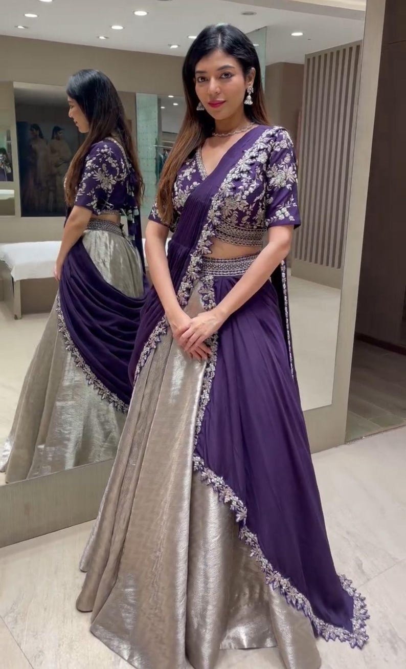 Puede incluir: Un conjunto de lehenga en color morado intenso y plata metalizada que incluye una blusa de manga corta con intrincados bordados plateados, una falda de talle alto a juego y un dupatta morado drapeado con un borde bordado en plata.