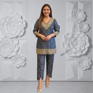 May include: A navy blue tunic and trousers set featuring a repeating white circular pattern. The tunic has three-quarter length sleeves and a V-neckline, both adorned with intricate gold embroidery. The hem also features a wide gold border.