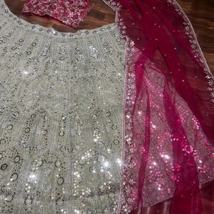 May include: A three-piece ensemble featuring a light sage green lehenga skirt, a matching sleeveless blouse, and a deep magenta dupatta. The skirt and blouse are heavily embellished with silver sequins and intricate embroidery. The sheer magenta dupatta features a wide, ornate silver border with sequins.