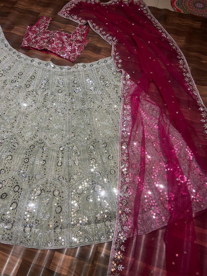May include: A three-piece ensemble featuring a light sage green lehenga skirt, a matching sleeveless blouse, and a deep magenta dupatta. The skirt and blouse are heavily embellished with silver sequins and intricate embroidery. The sheer magenta dupatta features a wide, ornate silver border with sequins.