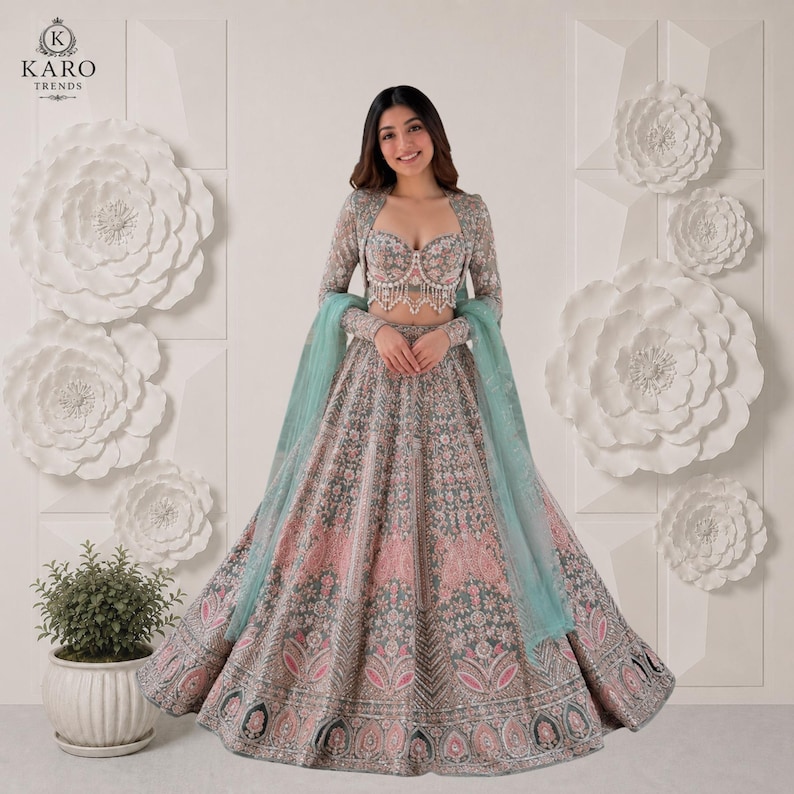 Puede incluir: Un conjunto de lehenga en verde salvia y rosa empolvado que incluye un top tipo bustier con escote coraz&oacute;n, adornado con cuentas y bordados, una falda larga acampanada a juego y un dupatta verde claro transparente sobre los brazos.