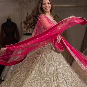 May include: A floor-length lehenga skirt featuring a gold hexagonal pattern with embedded mirror work, paired with a matching long-sleeved blouse and a vibrant hot pink sheer dupatta with an ornate gold embroidered border draped across the body.