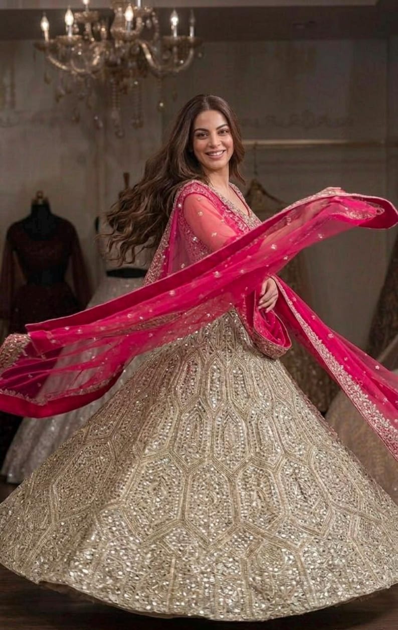 May include: A floor-length lehenga skirt featuring a gold hexagonal pattern with embedded mirror work, paired with a matching long-sleeved blouse and a vibrant hot pink sheer dupatta with an ornate gold embroidered border draped across the body.