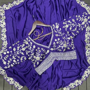 Puede incluir: Un conjunto de sari y blusa de color morado intenso con intrincados bordados florales plateados a lo largo de los bordes y las mangas. La blusa tiene un escote en V y un dobladillo estructurado, mientras que la tela del sari tiene un acabado satinado.