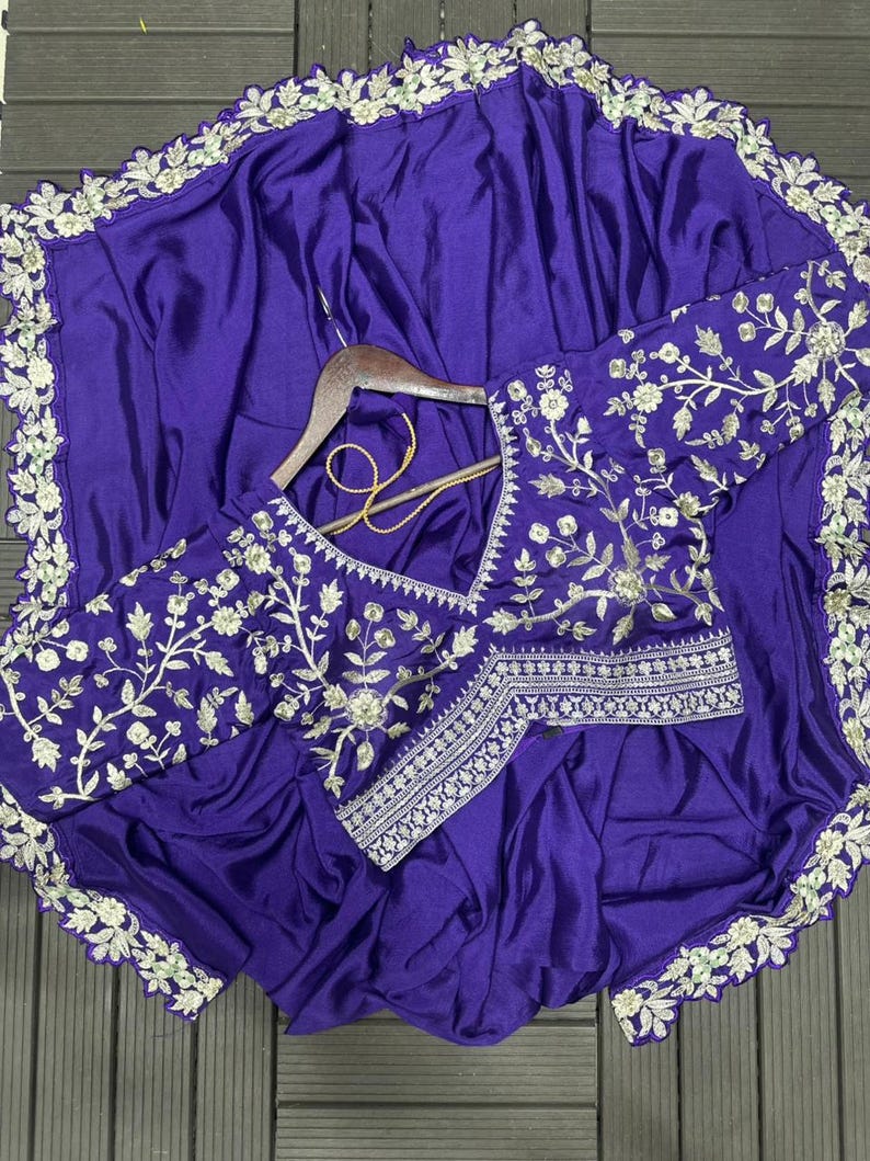 Puede incluir: Un conjunto de sari y blusa de color morado intenso con intrincados bordados florales plateados a lo largo de los bordes y las mangas. La blusa tiene un escote en V y un dobladillo estructurado, mientras que la tela del sari tiene un acabado satinado.