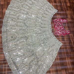May include: A light sage green lehenga skirt featuring an intricate pattern of silver embroidery and reflective mirror work, paired with a separate sleeveless blouse in a deep magenta pink, also adorned with matching silver embroidery and mirror details.