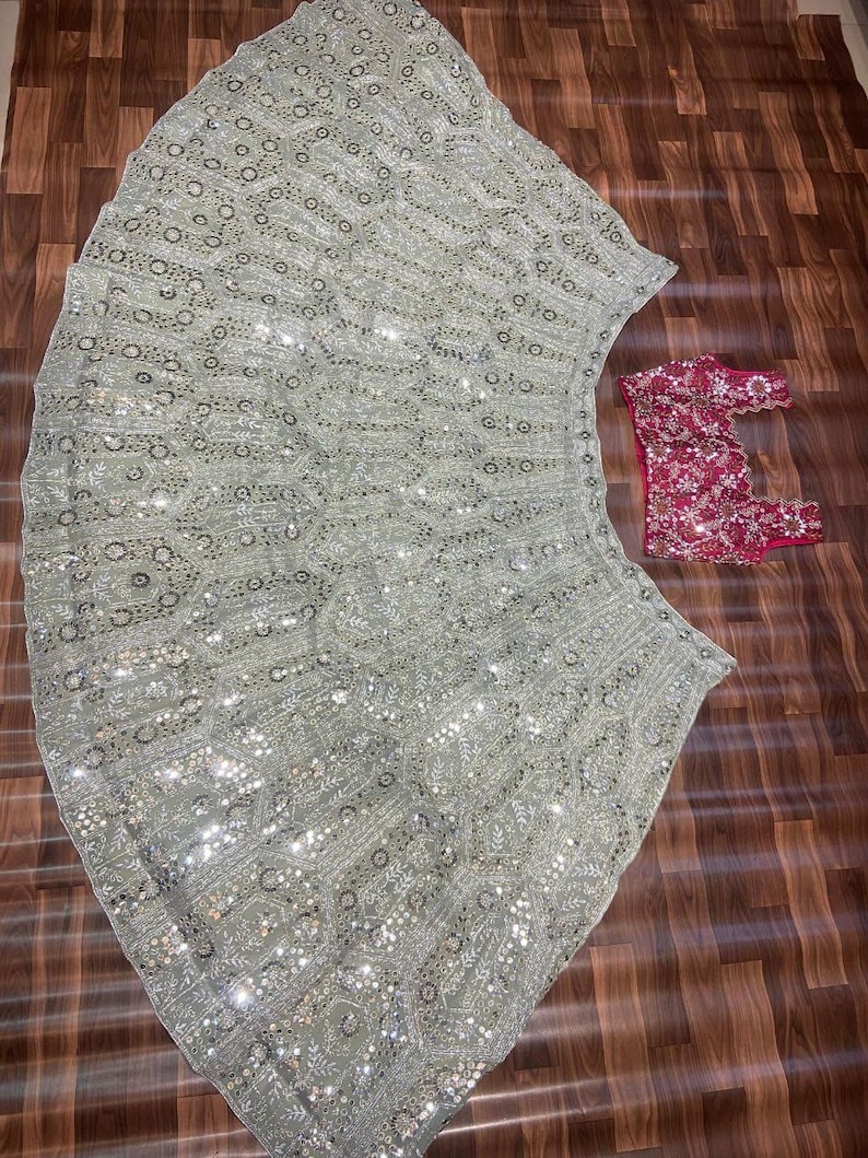 May include: A light sage green lehenga skirt featuring an intricate pattern of silver embroidery and reflective mirror work, paired with a separate sleeveless blouse in a deep magenta pink, also adorned with matching silver embroidery and mirror details.