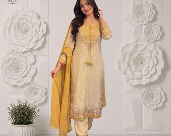 Puede incluir: Una túnica de color crema de manga larga con bordados dorados, un pantalón a juego y una dupatta de color amarillo mostaza. La túnica presenta un cuello redondo con borlas decorativas y detalles dorados intrincados en el dobladillo y el pecho.