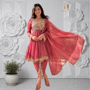 May include: A dusty rose-coloured peplum-style tunic with long sleeves, featuring intricate gold-toned mirror work embroidery along the neckline, cuffs, and hem. The outfit includes matching dhoti-style trousers and a sheer, flowing dupatta scarf.