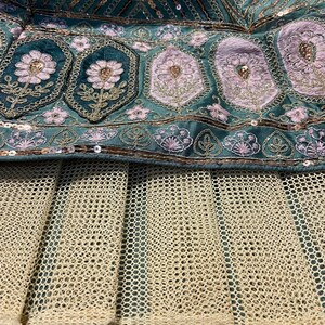 Puede incluir: Una prenda de tela color verde azulado con bordados intrincados de motivos florales rosas, hilo met&aacute;lico dorado y lentejuelas color cobre. Debajo de la secci&oacute;n bordada hay una capa de tela de malla beige con patr&oacute;n de rejilla y pliegues.