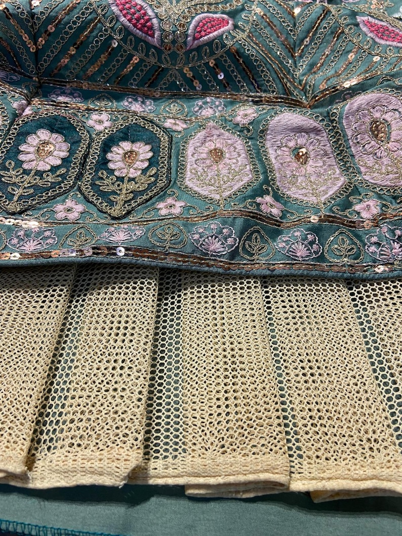Puede incluir: Una prenda de tela color verde azulado con bordados intrincados de motivos florales rosas, hilo met&aacute;lico dorado y lentejuelas color cobre. Debajo de la secci&oacute;n bordada hay una capa de tela de malla beige con patr&oacute;n de rejilla y pliegues.