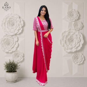 Pode incluir: Um vestido longo de cor fúcsia vibrante com mangas curtas, uma saia drapeada estilo envelope e um intrincado bordado branco ao longo do corpete, mangas e bainha. A peça inclui um cinto a condizer e apresenta uma silhueta fluida e elegante.