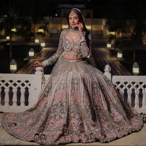 Puede incluir: Un conjunto de lehenga en color verde salvia y rosa empolvado con intrincados bordados plateados, abalorios y lentejuelas. El conjunto incluye una blusa corta de manga larga con escote coraz&oacute;n y una falda plisada voluminosa.