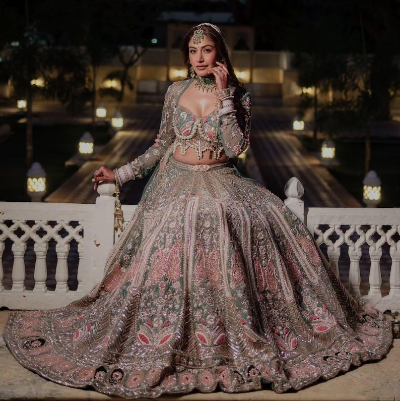 Puede incluir: Un conjunto de lehenga en color verde salvia y rosa empolvado con intrincados bordados plateados, abalorios y lentejuelas. El conjunto incluye una blusa corta de manga larga con escote coraz&oacute;n y una falda plisada voluminosa.