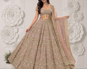 Puede incluir: Un conjunto de lehenga choli en color beige que incluye una blusa sin mangas y una falda voluminosa hasta el suelo, ambas decoradas con intrincados bordados dorados, lentejuelas y cuentas, junto a una dupatta transparente a juego.