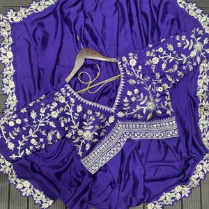 Puede incluir: Una prenda de color morado intenso con mangas largas y escote en V, adornada con intrincados bordados florales plateados. Los bordes de la tela est&aacute;n rematados con un borde festoneado de bordado plateado a juego, creando un dise&ntilde;o coherente.