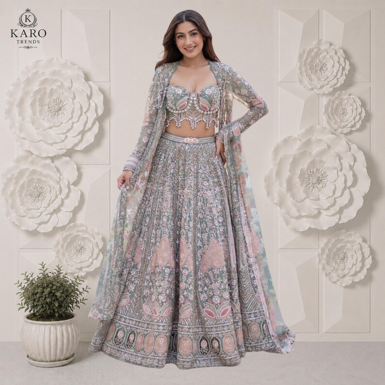 Puede incluir: Un conjunto lehenga verde salvia que presenta un corpi&ntilde;o sin tirantes con escote coraz&oacute;n y una falda larga hasta el suelo, ambos muy adornados con intrincados bordados plateados, lentejuelas y detalles florales en rosa suave, junto con una chaqueta transparente a juego.