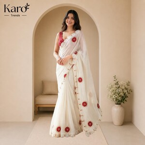 May include: A white saree featuring a sleeveless blouse with a red patterned design. The saree is adorned with circular red embroidered motifs along the border and the pallu. The text Karo Trends is visible in the top left corner of the image.