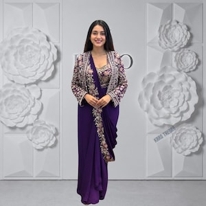 Puede incluir: Un vestido largo de color morado intenso con cuerpo sin mangas y falda drapeada. La prenda se combina con una chaqueta a juego de manga larga adornada con intrincados bordados florales en dorado y blanco a lo largo de los bordes y mangas.