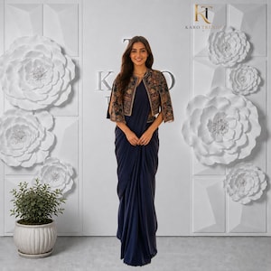 May include: A dark blue floor-length saree paired with a cropped jacket featuring intricate multi-coloured floral embroidery and short sleeves. The ensemble is worn against a white wall decorated with large paper flowers and the text KARO TRENDS.