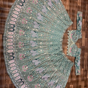 Puede incluir: Un conjunto de lehenga verde salvia que incluye una falda acampanada y una blusa de manga larga a juego. La tela est&aacute; densamente decorada con intrincados bordados plateados, motivos florales rosas, patrones de cachemira y peque&ntilde;os espejos reflectantes.