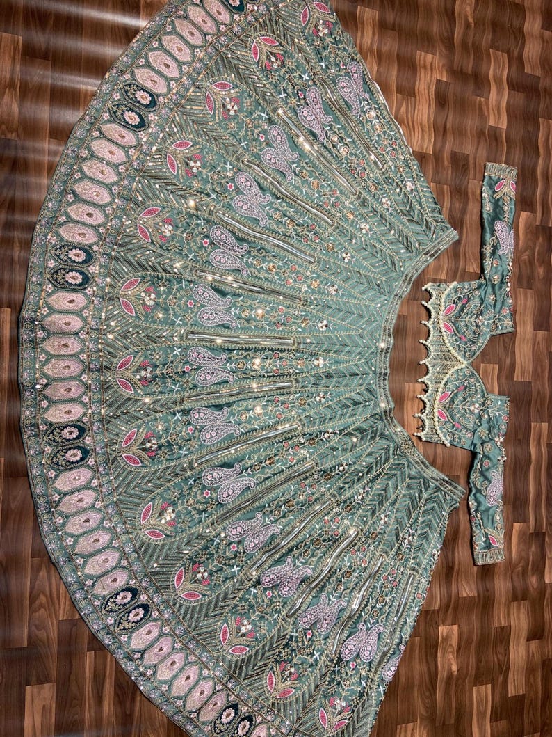 Puede incluir: Un conjunto de lehenga verde salvia que incluye una falda acampanada y una blusa de manga larga a juego. La tela est&aacute; densamente decorada con intrincados bordados plateados, motivos florales rosas, patrones de cachemira y peque&ntilde;os espejos reflectantes.