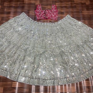 May include: A two-piece ensemble consisting of a sleeveless, square-neck magenta blouse with intricate gold embroidery and a voluminous, floor-length light gray skirt featuring dense silver sequin embellishments and detailed floral embroidery patterns.