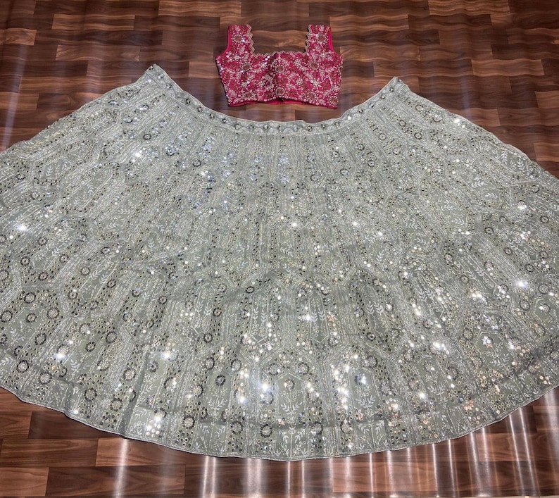 May include: A two-piece ensemble consisting of a sleeveless, square-neck magenta blouse with intricate gold embroidery and a voluminous, floor-length light gray skirt featuring dense silver sequin embellishments and detailed floral embroidery patterns.