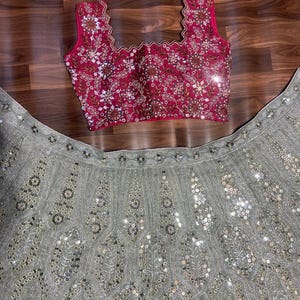 May include: A sleeveless fuchsia blouse with a scalloped neckline and intricate silver embroidery and mirror work, paired with a sage green skirt featuring elaborate silver thread embroidery and circular mirror embellishments throughout the fabric.