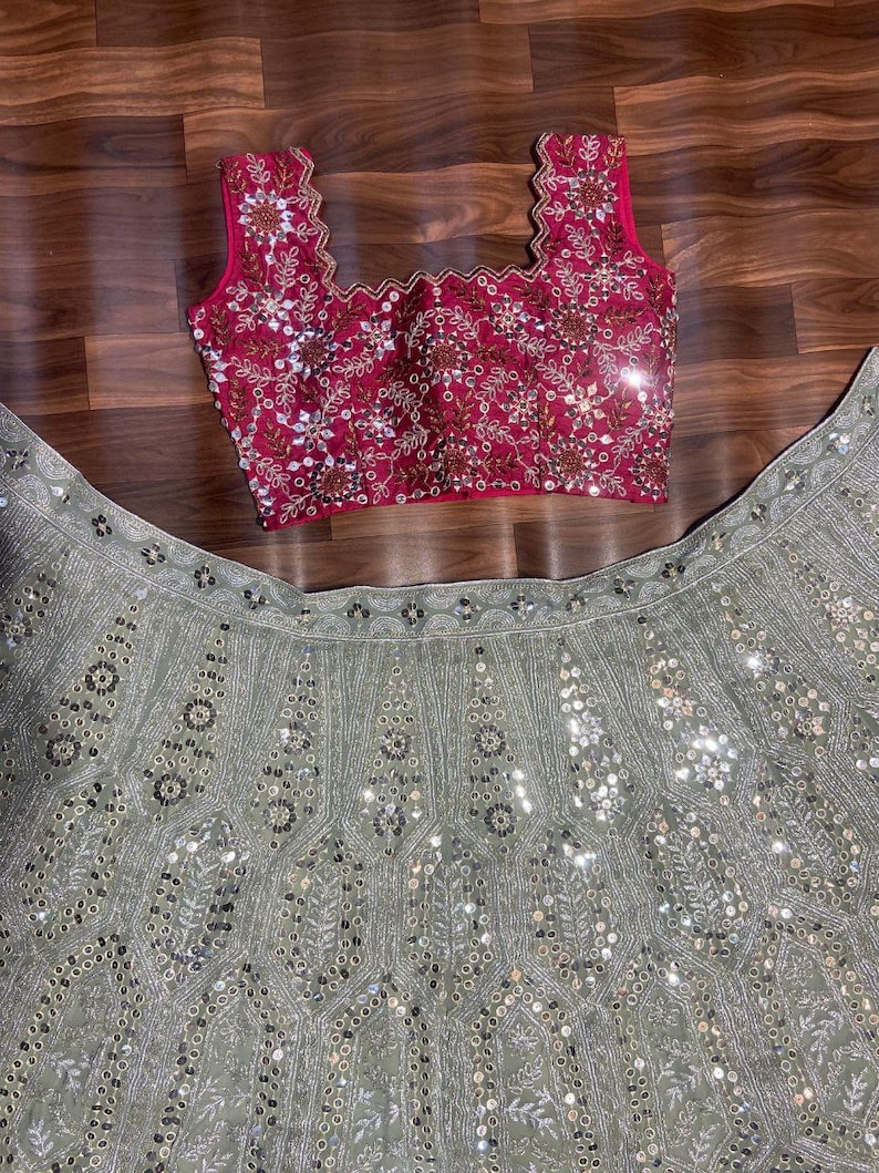 May include: A sleeveless fuchsia blouse with a scalloped neckline and intricate silver embroidery and mirror work, paired with a sage green skirt featuring elaborate silver thread embroidery and circular mirror embellishments throughout the fabric.