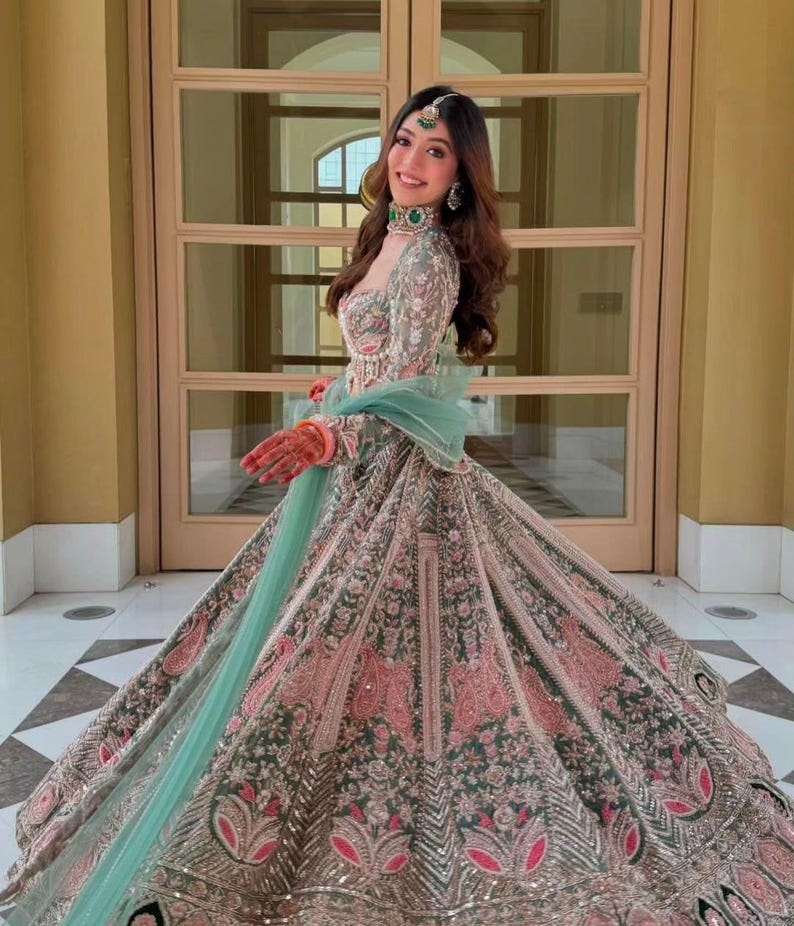 Puede incluir: Un lehenga choli largo hasta el suelo con bordados intrincados en tonos verde salvia, rosa empolvado y plata. El conjunto incluye un corpi&ntilde;o ajustado de manga larga, una falda voluminosa con motivos florales detallados y un dupatta verde menta.