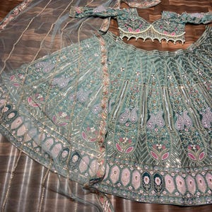 Puede incluir: Un conjunto lehenga verde salvia que incluye una blusa corta con mangas descubiertas y borde de perlas, una falda voluminosa con intrincados bordados florales y de cachemira, y un dupatta transparente a juego con detalles de lentejuelas.