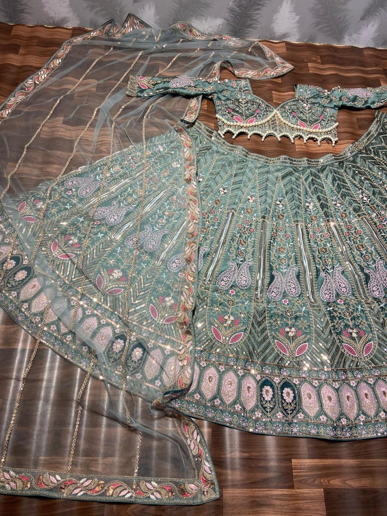 Puede incluir: Un conjunto lehenga verde salvia que incluye una blusa corta con mangas descubiertas y borde de perlas, una falda voluminosa con intrincados bordados florales y de cachemira, y un dupatta transparente a juego con detalles de lentejuelas.