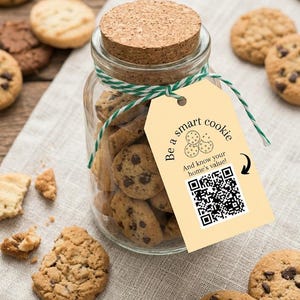 Op de afbeelding: Een doorzichtige glazen pot gevuld met koekjes met chocoladestukjes, afgesloten met een kurken deksel. Een lichtgeel rechthoekig label is met groen-wit touw aan de pot bevestigd. Het label bevat de tekst Be a smart cookie en een QR-code.