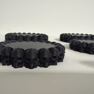 May include: A set of three circular black coasters, each featuring a flat top surface surrounded by a decorative border of small, detailed skull figures arranged in a continuous ring around the entire outer edge of the coaster.