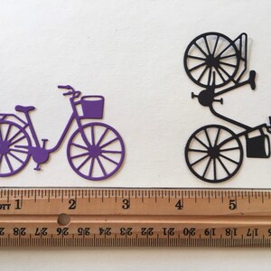 Puede incluir: Dos recortes de papel de bicicletas, uno morado y otro negro, cada uno con una cesta, se muestran sobre una superficie blanca. Una regla de madera con marcas en pulgadas se coloca debajo de los recortes.