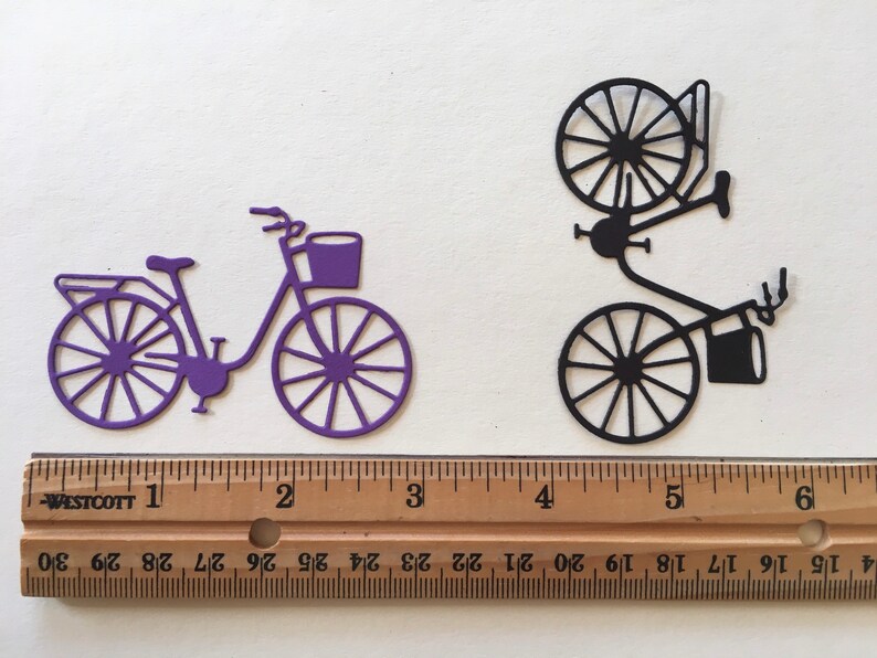 Puede incluir: Dos recortes de papel de bicicletas, uno morado y otro negro, cada uno con una cesta, se muestran sobre una superficie blanca. Una regla de madera con marcas en pulgadas se coloca debajo de los recortes.