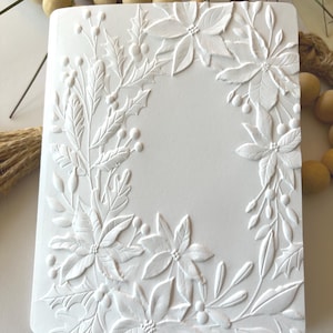 May include: A white, rectangular journal with a detailed embossed floral design. The design features poinsettias, holly, and berries, creating a festive, seasonal theme. The embossed elements add texture and depth to the cover.