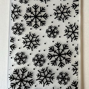May include: Clear, rectangular embossing folder featuring a repeating pattern of black snowflakes and small black dots. The snowflakes vary in size and are evenly spaced across the surface, creating a winter theme.
