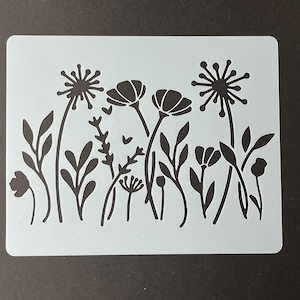 May include: A light blue stencil featuring a floral design with various flower and leaf silhouettes in black. The stencil is rectangular with rounded corners, suitable for crafts and DIY projects.